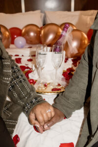 Couple holding hands for proposal photos with champagne on hotel bed