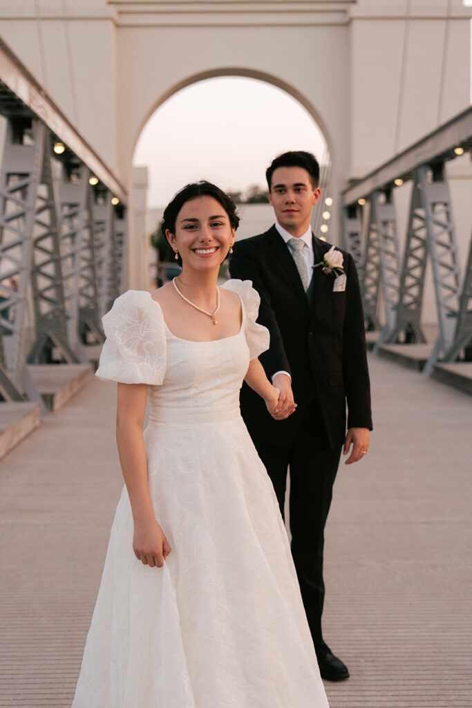 Bride and groom holding hands at suspension bridge during sunset bridal portraits after wedding